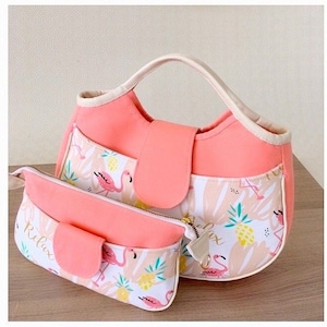 May include: A pink and white tote bag with a flamingo and pineapple print. The bag has a top handle and a zipper closure. It comes with a matching pink and white zippered pouch that says "Relax" on it.