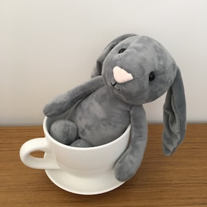 Puede incluir: Un conejito de peluche gris sentado en una taza de té y un platillo blancos.