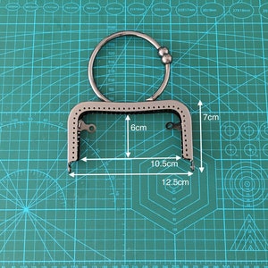 Puede incluir: Un marco de bolso de metal plateado con una abertura de 15,2 cm por 26,7 cm y una altura de 17,8 cm. El marco tiene un anillo decorativo con una cuenta en el extremo.