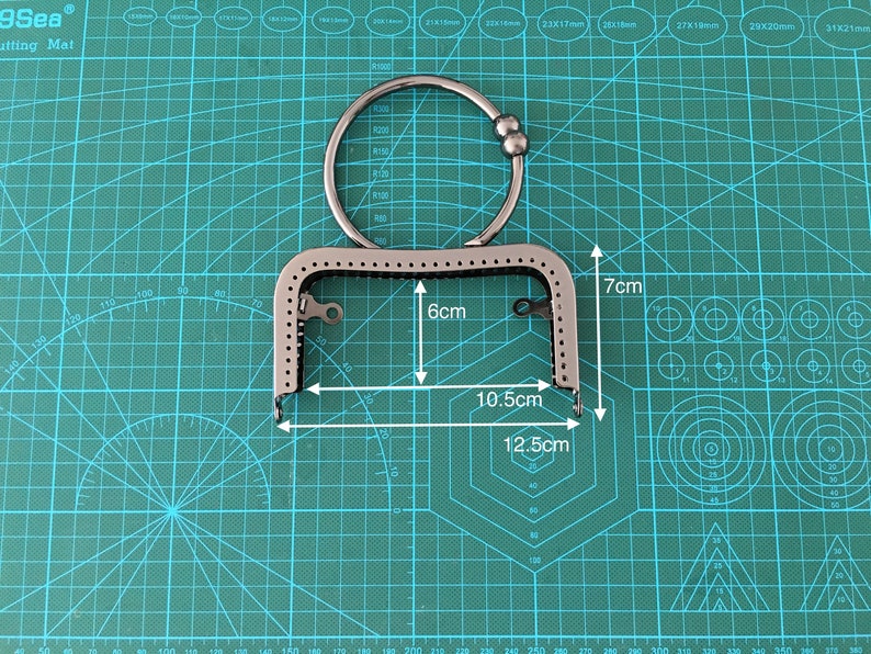 Puede incluir: Un marco de bolso de metal plateado con una abertura de 15,2 cm por 26,7 cm y una altura de 17,8 cm. El marco tiene un anillo decorativo con una cuenta en el extremo.