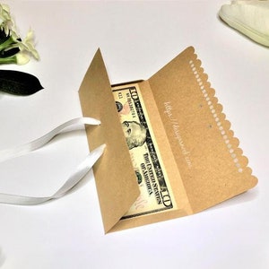 May include: A brown paper money envelope with a white satin ribbon. The envelope has a scalloped edge and a white label with the text "https://designpack.com". A US dollar bill is visible inside the envelope.