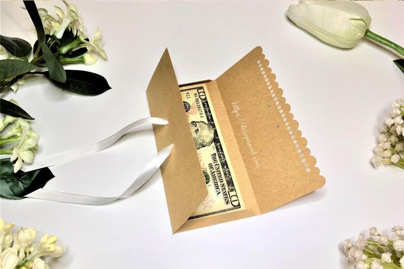 May include: A brown paper money envelope with a white satin ribbon. The envelope has a scalloped edge and a white label with the text "https://designpack.com". A US dollar bill is visible inside the envelope.