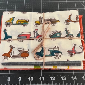 May include: A stack of four fabric squares with a white background and a repeating pattern of colourful scooters. The fabric squares are tied together with twine. The fabric squares are on a black and white grid surface.