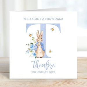 May include: A blue and white card with a watercolor illustration of Peter Rabbit standing in front of a blue letter T. The card says "Welcome to the world" and "Theodore" with the date "5th January 2022".