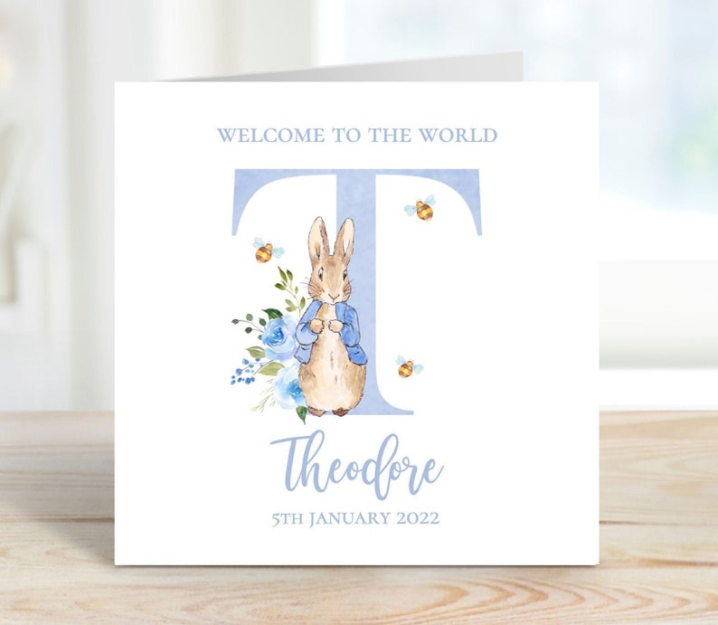 May include: A blue and white card with a watercolor illustration of Peter Rabbit standing in front of a blue letter T. The card says "Welcome to the world" and "Theodore" with the date "5th January 2022".