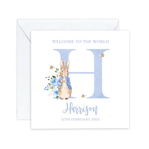 May include: A blue and white baby shower card with a watercolor illustration of Peter Rabbit in a blue jacket. The card features the letter H and the name Harrison with the date 12th February 2021. The text "Welcome to the World" is at the top of the card.
