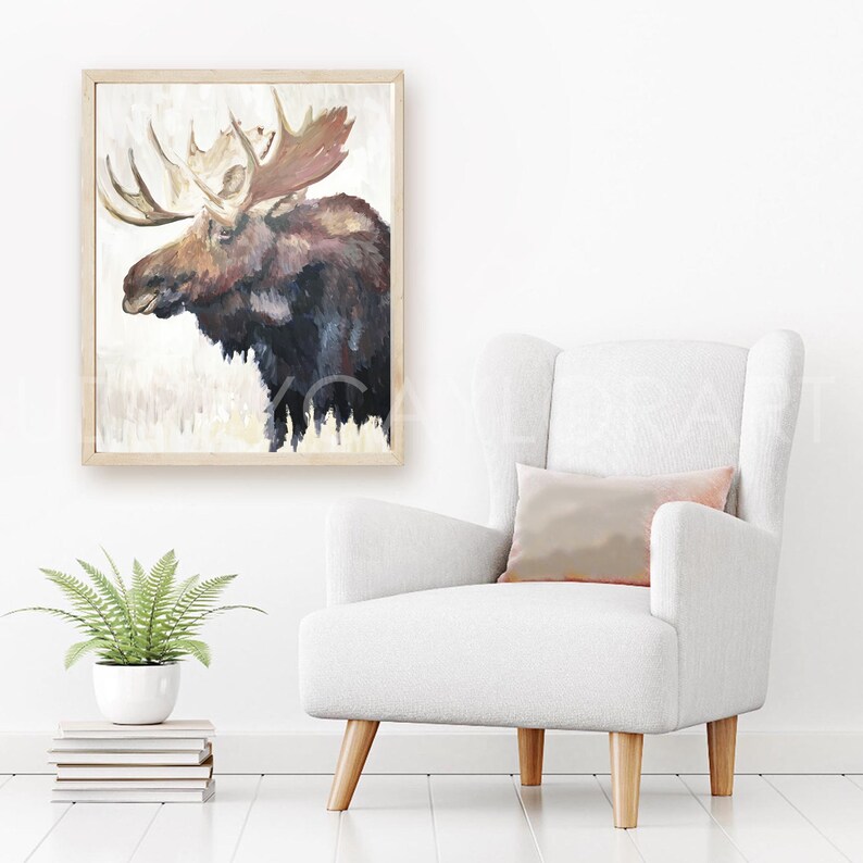 Peut inclure: Une peinture encadr&eacute;e d'un &eacute;lan brun et noir avec de grands bois. La peinture est accroch&eacute;e &agrave; un mur blanc au-dessus d'un fauteuil blanc avec un coussin rose clair.
