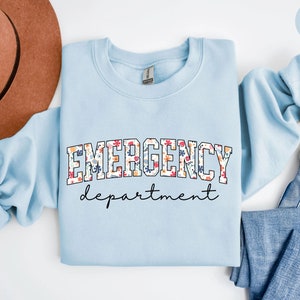 May include: Light blue sweatshirt with the text "EMERGENCY department" in black lettering with a floral pattern. The sweatshirt is folded and laid on a white surface with a brown hat and blue jeans.