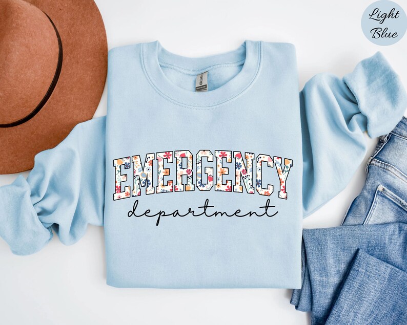 May include: Light blue sweatshirt with the text "EMERGENCY department" in black lettering with a floral pattern. The sweatshirt is folded and laid on a white surface with a brown hat and blue jeans.