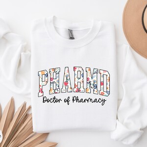 May include: White sweatshirt with the text "PHARMD Doctor of Pharmacy" in black lettering. The word "PHARMD" is decorated with a floral pattern in shades of pink, red, blue, yellow, and green.