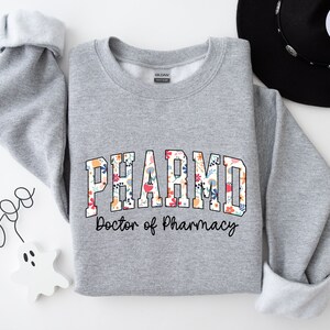 May include: A gray sweatshirt with a floral print design that says "PHARMD Doctor of Pharmacy".