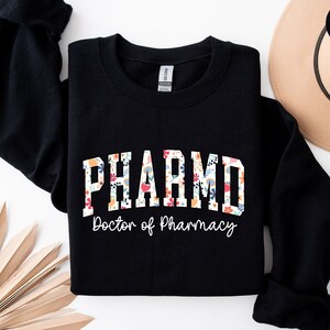 May include: Black sweatshirt with a floral design that says "PHARM.D. Doctor of Pharmacy"