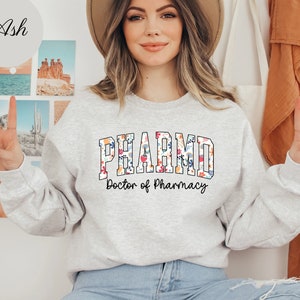 May include: A gray sweatshirt with a floral design that says "PHARM.D. Doctor of Pharmacy" in a collegiate font.