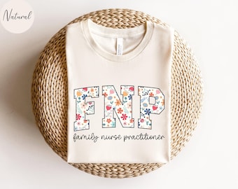 May include: Cream-colored t-shirt with the letters "FNP" in a floral design, with the words "family nurse practitioner" below. The shirt is displayed on a woven, circular surface.