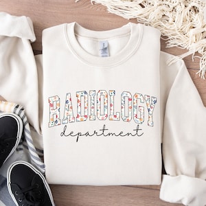 Op de afbeelding: Een wit sweatshirt met de tekst "RADIOLOGY department" in zwarte letters. Het woord "RADIOLOGY" is gevuld met een bloemenpatroon in roze, blauwe en gele tinten.