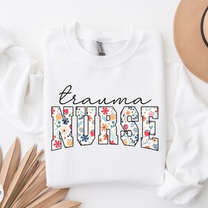 K&ouml;nnte beinhalten: Wei&szlig;er Pullover mit dem Text "trauma nurse" in schwarzen Buchstaben. Das Wort "nurse" ist in gro&szlig;en, fetten Buchstaben mit einem Blumenmuster.
