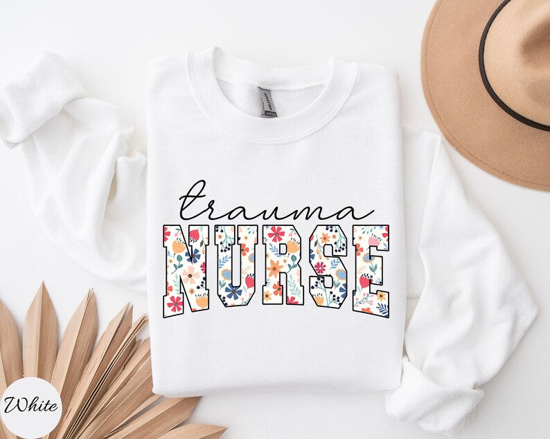 K&ouml;nnte beinhalten: Wei&szlig;er Pullover mit dem Text "trauma nurse" in schwarzen Buchstaben. Das Wort "nurse" ist in gro&szlig;en, fetten Buchstaben mit einem Blumenmuster.