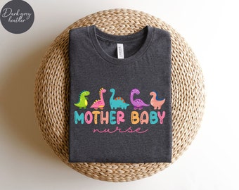 Może przedstawiać: Koszulka w kolorze ciemnoszary melanż z grafiką kolorowych dinozaurów i tekstem "Mother Baby Nurse".