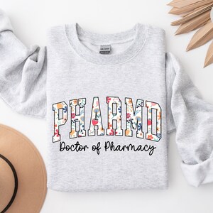 May include: A gray sweatshirt with the text "PHARM.D. Doctor of Pharmacy" in a floral font. The text is in a collegiate style.