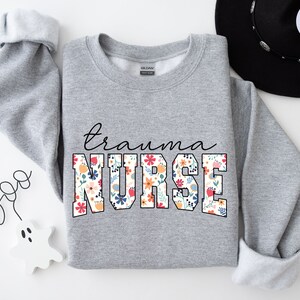K&ouml;nnte beinhalten: Ein grauer Pullover mit dem Text "trauma nurse" in gro&szlig;en wei&szlig;en Buchstaben mit einem Blumenmuster. Die Buchstaben sind schwarz umrandet.