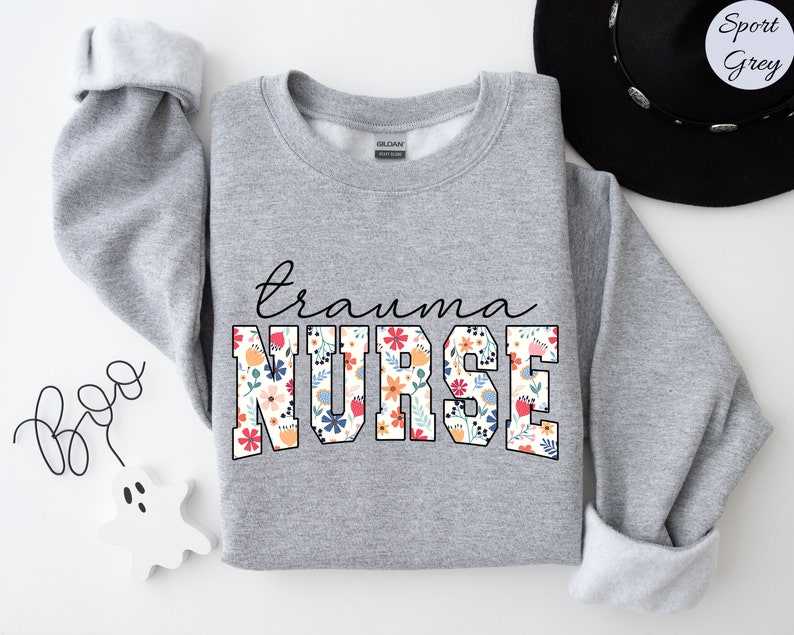 K&ouml;nnte beinhalten: Ein grauer Pullover mit dem Text "trauma nurse" in gro&szlig;en wei&szlig;en Buchstaben mit einem Blumenmuster. Die Buchstaben sind schwarz umrandet.