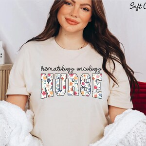 Puede incluir: Una camiseta de color crema con el texto "hematology oncology NURSE" en negro con detalles florales.