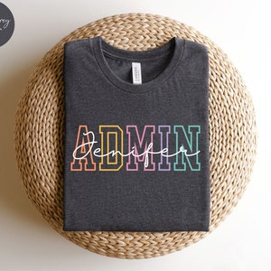 May include: Dark grey heather t-shirt with the word "ADMIN" in colorful block letters and the name "Jennifer" written in cursive underneath.