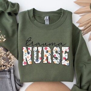 K&ouml;nnte beinhalten: Ein olivgr&uuml;ner Sweatshirt mit dem Text "trauma nurse" in Wei&szlig;, umrandet mit Schwarz. Die Buchstaben sind mit einem floralen Muster in den Farben Rosa, Rot, Gelb, Blau und Gr&uuml;n gef&uuml;llt.