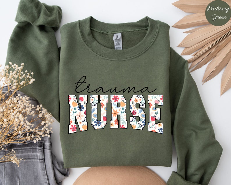 K&ouml;nnte beinhalten: Ein olivgr&uuml;ner Sweatshirt mit dem Text "trauma nurse" in Wei&szlig;, umrandet mit Schwarz. Die Buchstaben sind mit einem floralen Muster in den Farben Rosa, Rot, Gelb, Blau und Gr&uuml;n gef&uuml;llt.