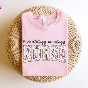 Puede incluir: Camiseta rosa con un dise&ntilde;o floral que dice "hematolog&iacute;a oncolog&iacute;a enfermera" en texto negro.
