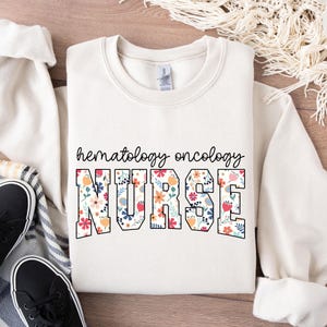 Może przedstawiać: Biała bluza z nadrukiem "hematology oncology NURSE" w czarnych obramowanych literach z kwiatowym wzorem wewnątrz liter. Bluza leży na drewnianej powierzchni z białym kocem i parą czarnych butów.