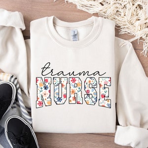 K&ouml;nnte beinhalten: Ein wei&szlig;er Pullover mit einem floralen Druckdesign. Der Text "trauma nurse" ist in Schwarz auf der Vorderseite des Pullovers gedruckt.