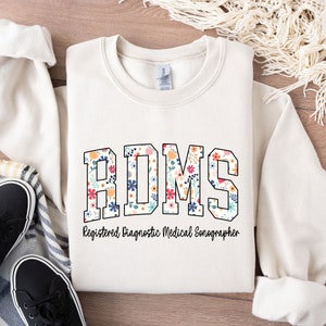 Könnte beinhalten: Ein weißer Pullover mit einem Blumendesign, das "RDMS" buchstabiert und den Text "Registered Diagnostic Medical Sonographer" trägt.