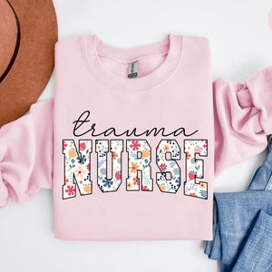 K&ouml;nnte beinhalten: Hellrosafarbener Pullover mit dem Text "trauma nurse" in schwarzen umrandeten Buchstaben. Die Buchstaben sind mit einem floralen Muster in den Farben Rosa, Rot, Gelb, Blau und Gr&uuml;n gef&uuml;llt.
