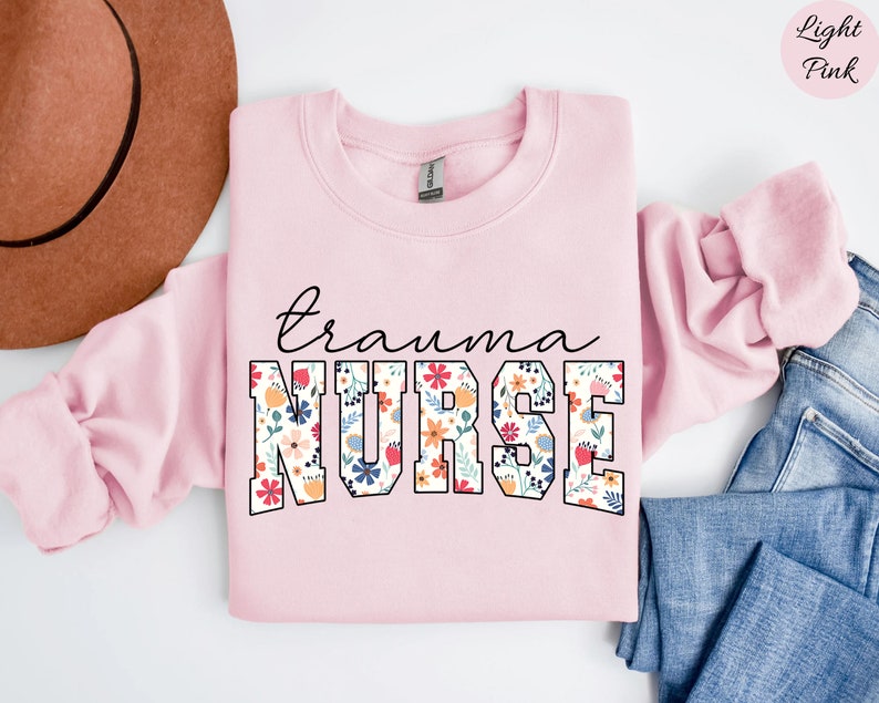 K&ouml;nnte beinhalten: Hellrosafarbener Pullover mit dem Text "trauma nurse" in schwarzen umrandeten Buchstaben. Die Buchstaben sind mit einem floralen Muster in den Farben Rosa, Rot, Gelb, Blau und Gr&uuml;n gef&uuml;llt.