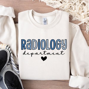 Könnte beinhalten: Ein weißer Pullover mit dem Text "RADIOLOGY department" in blauer und schwarzer Schrift mit einem schwarzen Herzen unter dem Text.
