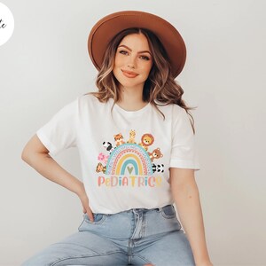 May include: White t-shirt with a rainbow and animals on it. The text "PEDIATRICS" is below the rainbow. The model is wearing a brown hat and blue jeans.