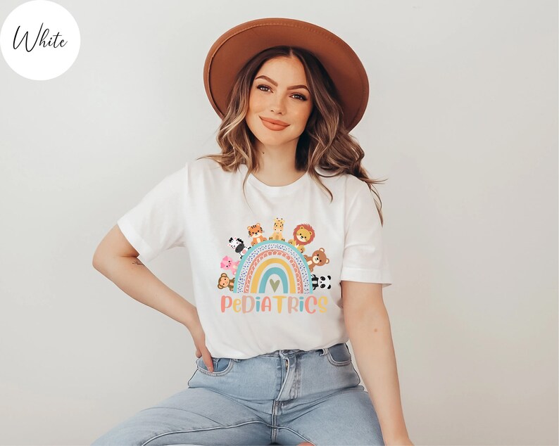 May include: White t-shirt with a rainbow and animals on it. The text "PEDIATRICS" is below the rainbow. The model is wearing a brown hat and blue jeans.