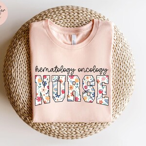Puede incluir: Una camiseta rosa con un dise&ntilde;o floral que dice "hematolog&iacute;a oncolog&iacute;a enfermera" en texto negro.