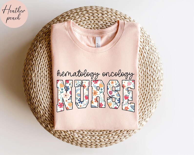 Puede incluir: Una camiseta rosa con un dise&ntilde;o floral que dice "hematolog&iacute;a oncolog&iacute;a enfermera" en texto negro.