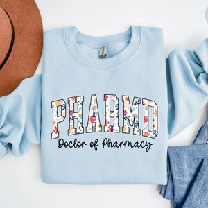 May include: Light blue sweatshirt with a floral print design that says "PHARM.D. Doctor of Pharmacy" in black text.