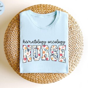 Puede incluir: Una camiseta azul claro con las palabras "hematology oncology nurse" en letras florales. La camiseta se muestra sobre una superficie de paja tejida.