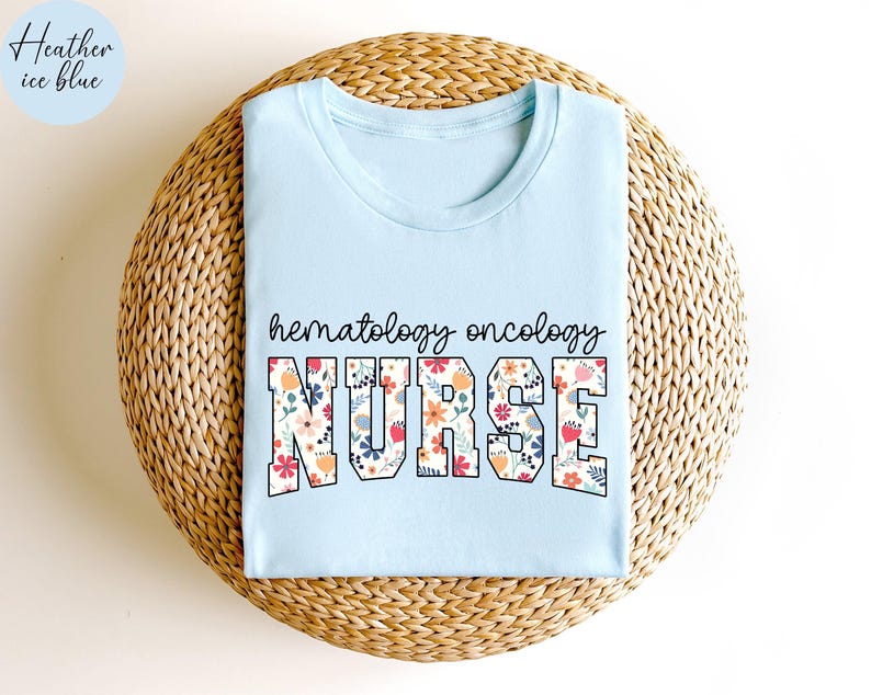Puede incluir: Una camiseta azul claro con las palabras "hematology oncology nurse" en letras florales. La camiseta se muestra sobre una superficie de paja tejida.