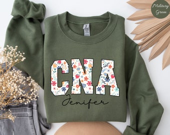 Op de afbeelding: Een groen sweatshirt met de letters "CNA" in wit met een bloemenpatroon in de letters. De naam "Jenifer" staat in cursief onder de letters.