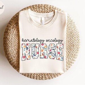 Puede incluir: Una camiseta blanca con un dise&ntilde;o floral que dice "hematology oncology nurse".