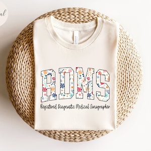 Könnte beinhalten: Ein naturfarbenes T-Shirt mit einem floralen Design, das "RDMS" buchstabiert und den Text "Registered Diagnostic Medical Sonographer" trägt.