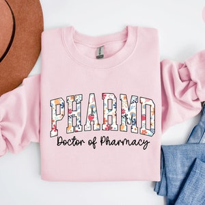 May include: Light pink sweatshirt with the text "PHARM.D. Doctor of Pharmacy" in black lettering with a floral pattern inside the letters.