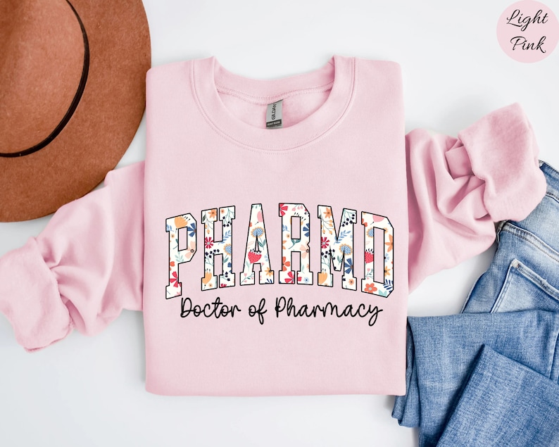 May include: Light pink sweatshirt with the text "PHARM.D. Doctor of Pharmacy" in black lettering with a floral pattern inside the letters.