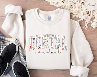 Puede incluir: Una sudadera blanca con un diseño de estampado floral que dice "DENTAL assistant" en texto negro.