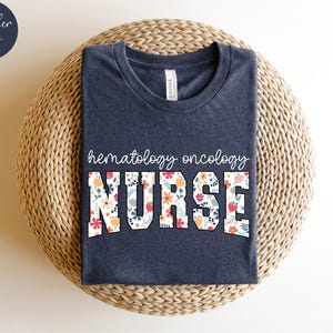 Puede incluir: Una camiseta azul marino jaspeada con el texto "hematolog&iacute;a oncolog&iacute;a ENFERMERA" en letras blancas con un patr&oacute;n floral.